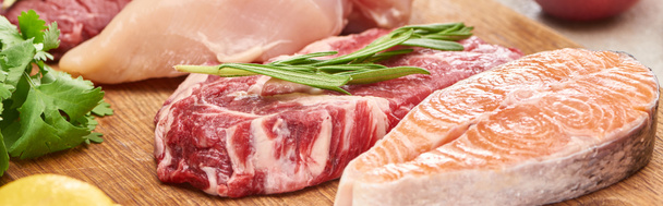 Tiro panorâmico de carne crua com galho de alecrim perto de salmão e frango com vegetação em tábua de corte de madeira
 - Foto, Imagem