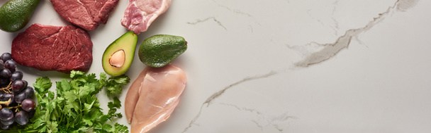 Panoramic shot of raw meat, chicken fillet, avocados, greenery and grape on marble surface with copy space - Photo, Image