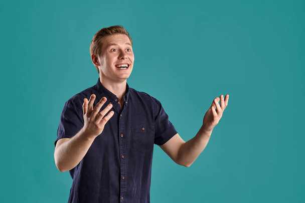 Close-up portrait of a young good-looking ginger peson in a stylish navy t-shirt looking wondered while posing on blue studio background. Human facial expressions. Sincere emotions concept. Copy space. - Photo, Image