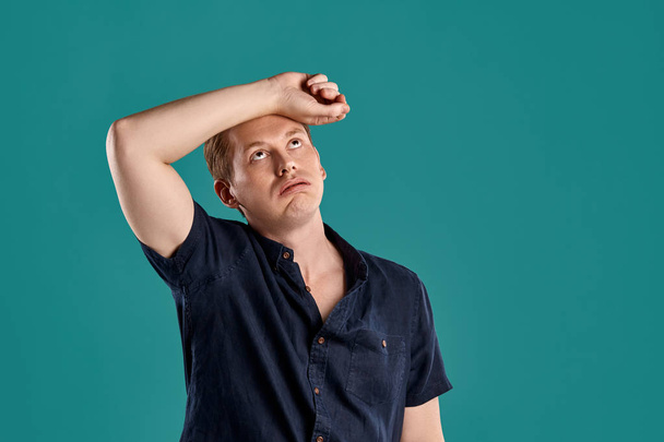 Close-up portrait of a young athletic ginger peson in a stylish navy t-shirt looking tired while posing on blue studio background. Human facial expressions. Sincere emotions concept. Copy space. - Photo, Image