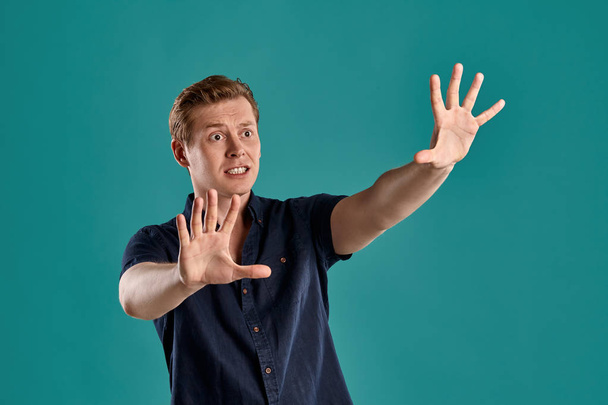 Close-up portrait of a young lovely ginger fellow in a stylish navy t-shirt looking scared and acting like stopping someone while posing on blue studio background. Human facial expressions. Sincere emotions concept. Copy space. - Φωτογραφία, εικόνα
