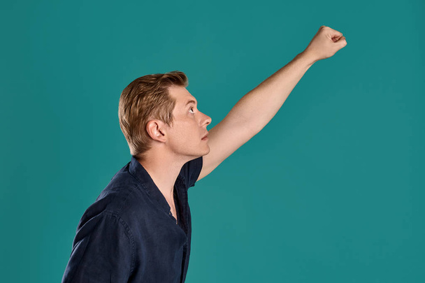 Close-up portrait of a young goodly ginger fellow in a stylish navy t-shirt acting like he is a superman while posing on blue studio background. Human facial expressions. Sincere emotions concept. Copy space. - Photo, Image