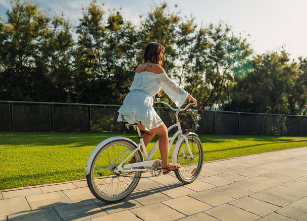 Asian model in a blue dress rides a white bicycle in a green park - Photo, Image