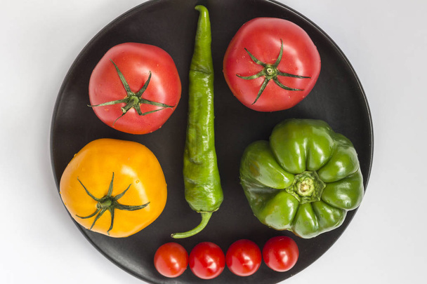 Still life of vegetables: tomatoes, peppers and cucumber in a black plate. White background. Top view - Photo, image