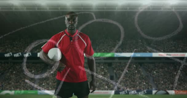Animation of an African American male rugby player standing, holding a ball under his arm and looking to camera with hand drawn game plan in the foreground and sports stadium in the background 4k - Imágenes, Vídeo