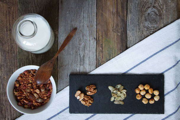 Tazón de granola crujiente de miel con semillas de lino, arándanos, una botella de leche y montones de nueces en un soporte negro en una mesa, enfoque selectivo
 - Foto, Imagen