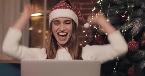 Young Beautiful Women Wearing Christmas Hat Sitting on the Sofa at Home Background Using her Laptop Rejoices Smiles and Laughs Clapes her Hands Concept of Holidays and New Year - Footage, Video