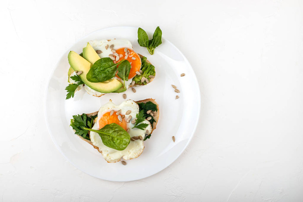 Fried eggs on a toasts bread with avocado, spinach and seeds on a white plate on the white background. - Foto, Bild