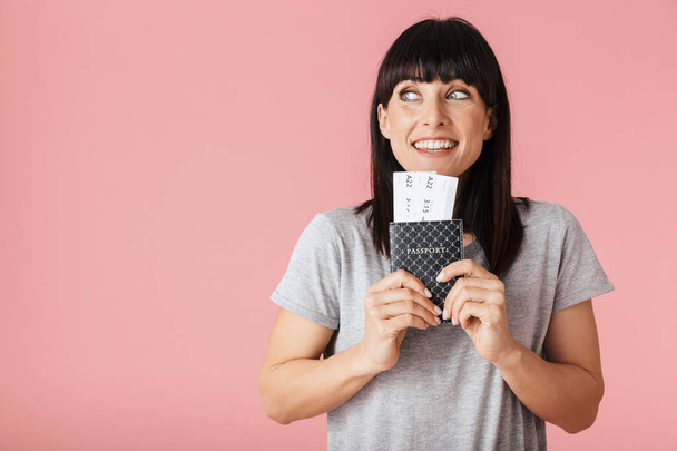 Excited happy woman posing isolated over light pink background wall holding passport with tickets. - Foto, afbeelding