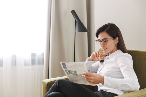 Concentrated beautiful young business woman in formal wear clothes indoors at home reading newspaper. - Photo, Image