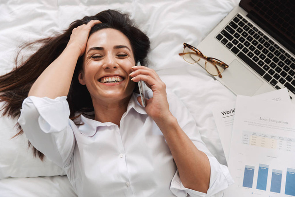Amazing beautiful happy young business woman in formal wear clothes indoors at home lies on bed talking by phone. - Photo, image