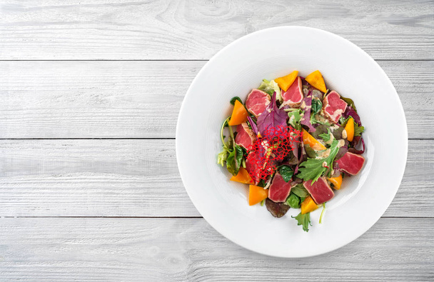 Healthy salad with seared tuna fish, greens, bell pepper and sauce in plate on light wooden background. Healthy food, clean eating, dieting, top view - Photo, image
