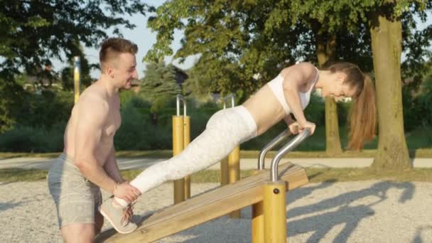 Αργή κίνηση, κοντινό πλάνο: Μυώδης προπονητής κρατώντας γυναίκα από τους αστραγάλους, ενώ κάνει push ups σχετικά με τον εξοπλισμό γυμναστικής πάρκο. Η χαρούμενη κοπέλα της κάνει πους απς σε ξύλινο παγκάκι ενώ ο φίλος της την επευφημεί.. - Πλάνα, βίντεο