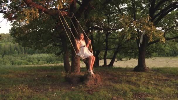jong meisje swingende op een schommel onder een boom in de zon, spelen met kinderen. Close-up. Familie plezier in de natuur. kind rijdt een touwschommel op een eiken tak in het Park de zonsondergang. meisje lacht, verheugt. - Video