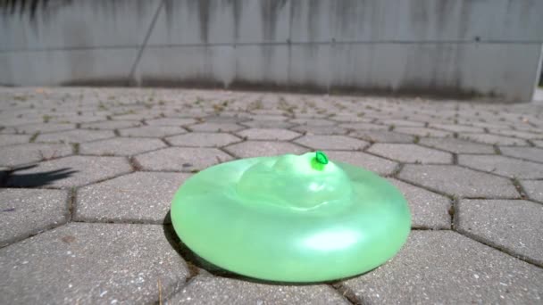 SLOW MOTION CLOSE UP: Green rubber balloon doesn 't pop after falling on the tiled ground. Красочная водяная бомба отскакивает от бетонной плитки после падения с неба. Зеленый шар наполнен водой
 - Кадры, видео