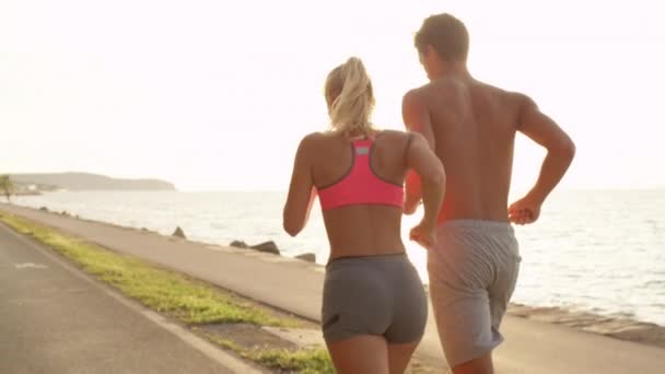 SLOW MOTION, LENS FLARE, CLOSE UP: Blonde woman and her boyfriend jog along the empty asphalt road next to the sea. Fit Caucasian couple jogging on a sunny summer day. Two athletic training partners. - Footage, Video