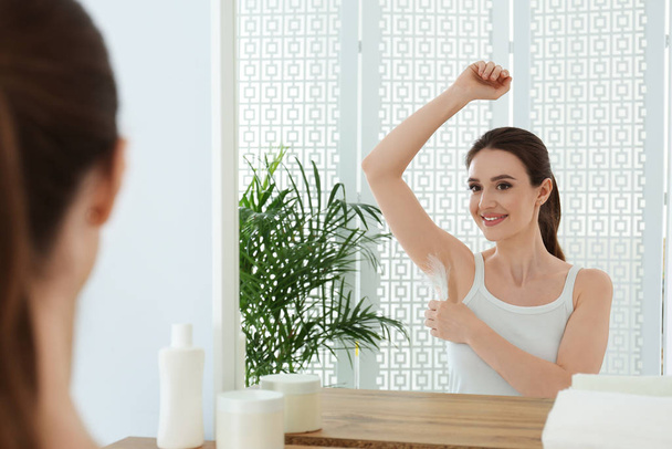 Young woman touching armpit with feather near mirror at home. Epilation procedure - Φωτογραφία, εικόνα