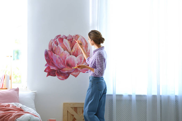 Young interior designer painting flower on wall in modern room - Foto, Imagen