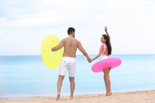 Happy young couple having fun with inflatable rings on beach near sea - Photo, image