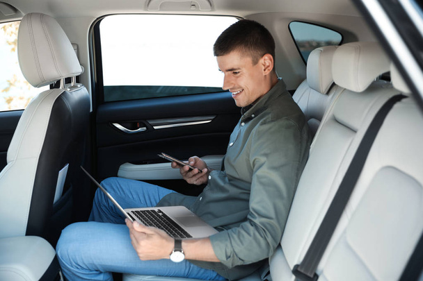 Attractive young man working with laptop and smartphone on backseat in luxury car - Fotografie, Obrázek