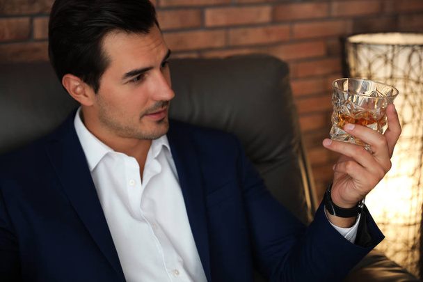 Young man with glass of whiskey near brick wall indoors - Φωτογραφία, εικόνα