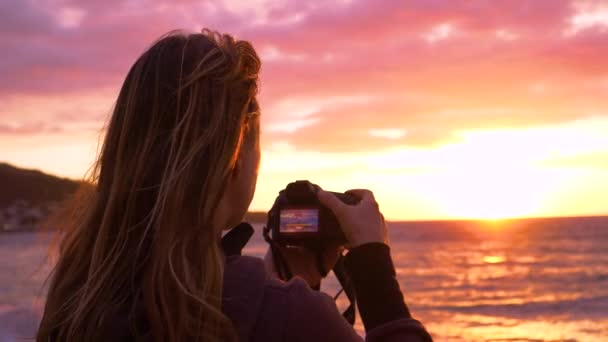 Молода жінка-фотограф захоплює мальовниче літнє вечірнє сонце над океаном. Ніжний бриз дме крізь волосся дівчат, поки вона фотографує мальовничий захід сонця
. - Кадри, відео