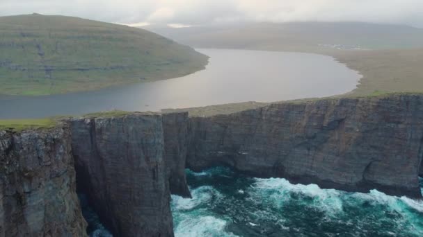 AEREO: Piccoli turisti seduti su una scogliera erbosa a guardare il maestoso paesaggio roccioso e i mari agitati in una giornata grigia nuvolosa nelle Isole Faroe. Due escursionisti irriconoscibili che osservano la natura
. - Filmati, video