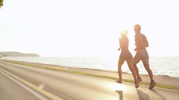 Copy Space, Lens Flare: Złote letnie promienie słoneczne świecą na sportową parę ćwiczącą razem nad morzem. Zdjęcia energicznego mężczyzny i kobiety biegających piękną nadmorską drogą w słoneczny dzień. - Materiał filmowy, wideo
