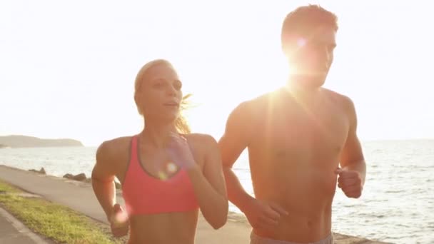 SLOW MOTION, LENS FLARE, CLOSE UP: Two cheerful young training partners jogging along the scenic ocean road. Cinematic shot of smiling athletic woman and man running on a beautiful sunny summer day. - Footage, Video