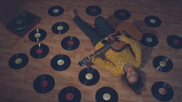 レコードの中でギターを持つ女の子。その少女は床に横たわっている。彼女の隣には、古いレコードとターンテーブルがたくさんあります。彼女はアコースティックギターを弾く. - 映像、動画