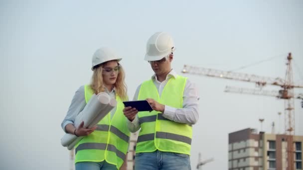 Dva průmysloví inženýrovi nosí bezpečnostní přilbu a drží v práci tabletové inženýrství a hovoří s prohlídkou výkresů. Na budovu venku. Inženýrské nástroje - Záběry, video