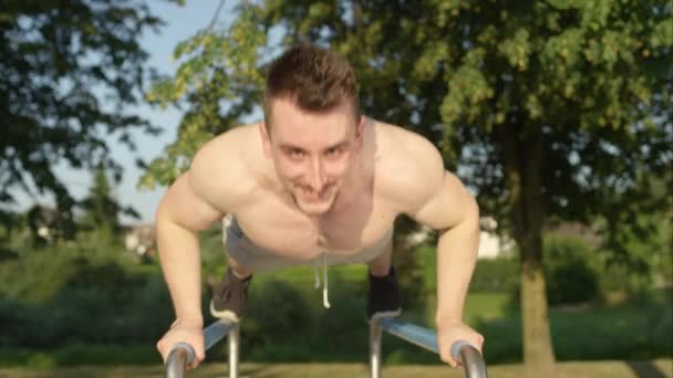 MOTION LENT, PORTRAIT : Un homme caucasien souriant fait des pompes de poids corporel sur l'équipement de gymnastique extérieur frais par une journée ensoleillée. Joyeux jeune athlète masculin faisant de l'exercice dans le parc appuie sur des barres parallèles
. - Séquence, vidéo