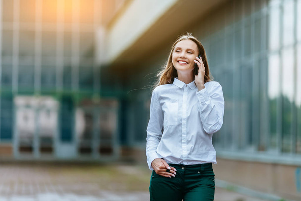 Young famale calling on a smartphone. Attractive businesswoman talking by phone. Office building Background. Technology internet and happy people concept. - 写真・画像