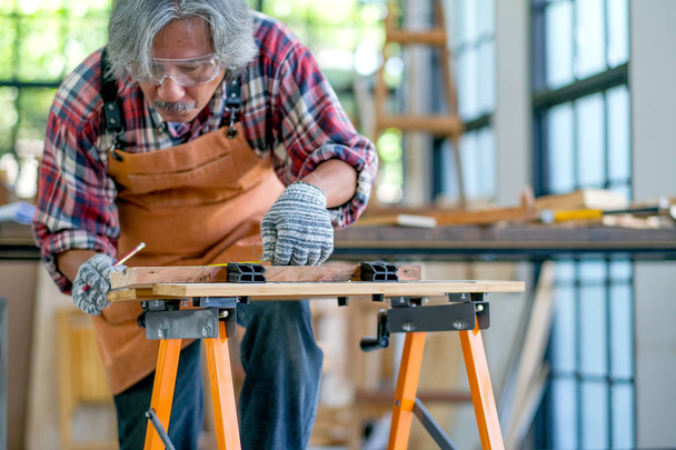 Starý asijský řemeslník působí jako měření dřeva v místnosti s hromadou dřeva a obklopené sklenicemi. - Fotografie, Obrázek