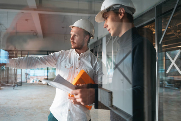 Building inspector hold blueprint in hand and have meeting with worker wearing safety hard hat on industrial construction site. Architect and build engineer discuss project drawing - Valokuva, kuva