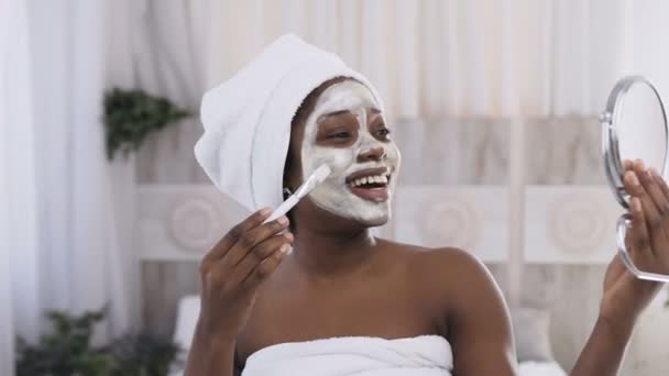 Happy African american woman in white towel on head applying yogurt mask on face, watching in the mirror. Healthy facial, scincare and rejuvenation concept - 映像、動画