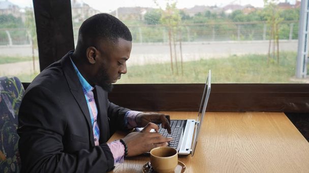 Czarny biznesmen pracuje wpisując wiadomość na laptopa siedzi w letniej kawiarni. - Zdjęcie, obraz