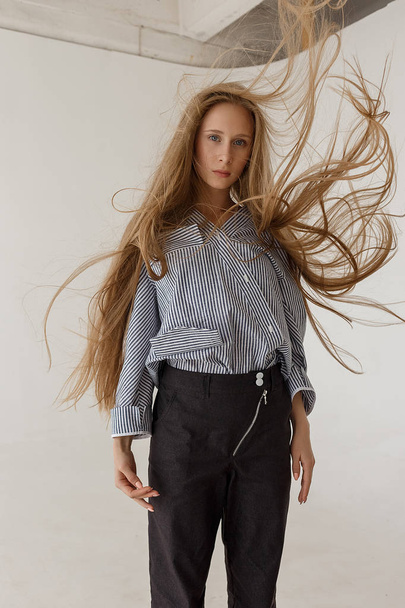 young teenage girl stands in front of white cyclorama in black jeans and striped oversize shirt with her hair fluttering - Fotó, kép