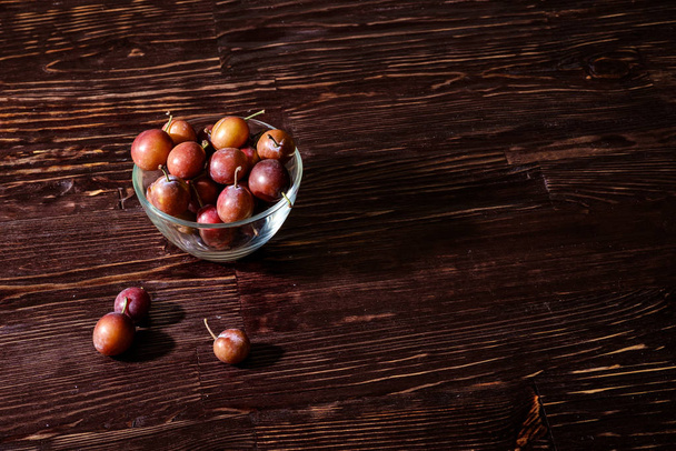 Zralý sladký švestkový plody ve skleněné misce poblíž roztroušených švestek na tmavém dřevěném stolku, tvrdé světlo, kopírovací prostor - Fotografie, Obrázek