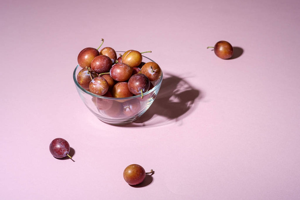 Ripe sweet plum fruits in glass bowl near with scattered plums on pink background, hard light, copy space - Photo, Image
