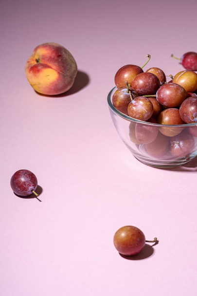Ripe sweet plum fruits in glass bowl near with scattered plums and peaches on pink background, hard light  - Photo, Image