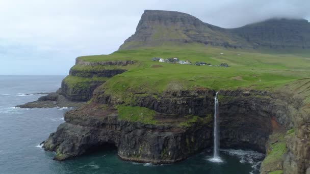 AERIAL: Picturesque village under grassy mountains and the spectacular cliffs on a cloudy day in the breathtaking Faroe Islands. Beautiful view of a spectacular waterfall in Scandinavian countryside. - Footage, Video