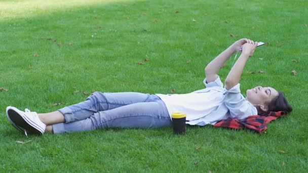 Young asian woman lying on the green grass. Relaxing, holding smartphone and listening to music in headphones. - 映像、動画