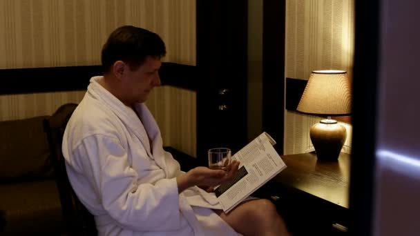 mężczyzna w białym płaszczu, siedząc przy stole z kieliszek whisky, czytając czasopismo - Materiał filmowy, wideo