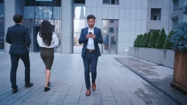 Młody człowiek biznesu używa smartphone podczas spaceru na Big City Business District Street. Klasycznie ubrany. Duży biurowy budynek w tle. Szukam sukcesu, pewny siebie. - Materiał filmowy, wideo