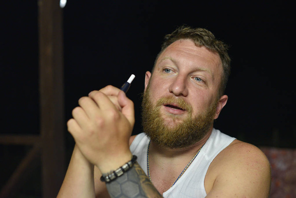 Ένας άντρας με κόκκινο μούσι καπνίζει ένα ηλεκτρονικό τσιγάρο.. - Φωτογραφία, εικόνα