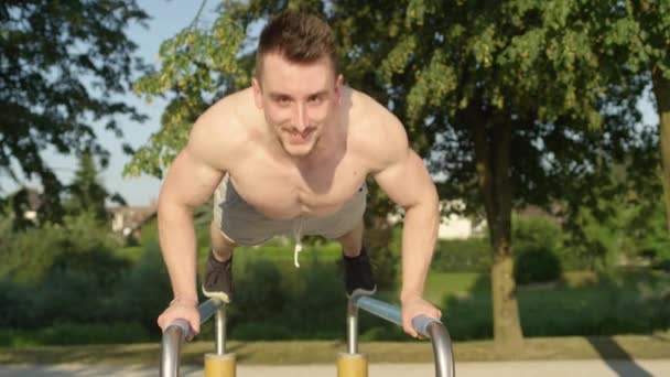 Pomalý pohyb, Portrét: Atletický mladý běloch dělá kliky na paralelních tyčích v zeleném parku. Usmívající se mužský atlet se baví cvičením ve venkovní tělocvičně v létě. Svalnatý chlap cvičení. - Záběry, video