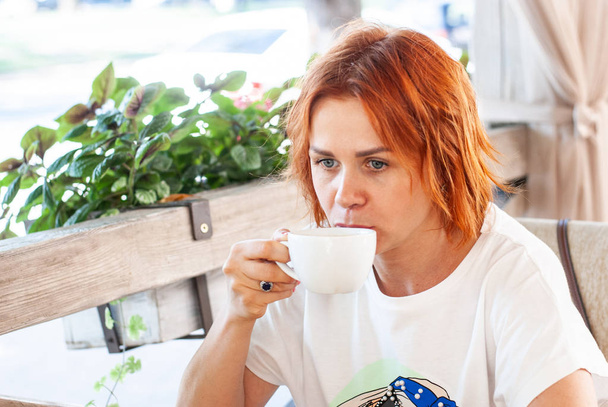 Ένα νεαρό κορίτσι με κόκκινα μαλλιά πίνει μαύρο καφέ από ένα λευκό φλιτζάνι  - Φωτογραφία, εικόνα
