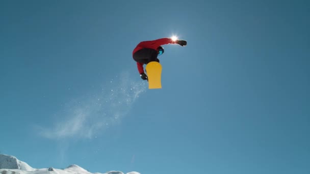 Slow Motion Close Up: Extreme snowboarder springt over de helderblauwe hemel in de zonnige bergwildernis. Freestyle boarder snowboarden en vliegen over camera in het skigebied. Snowboardsprong - Video