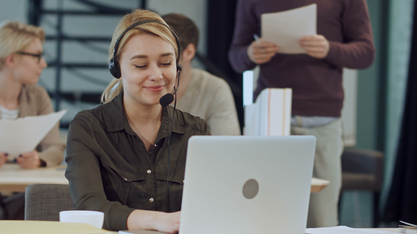Smiling female helpline operator with headphones at her desk in the office - Materiał filmowy, wideo